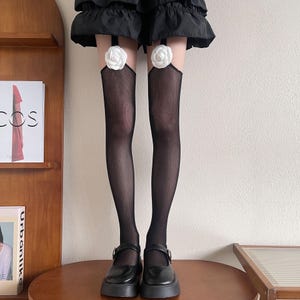 Puede incluir: Medias de rejilla negras con detalles de rosas blancas en la parte superior. Las medias se combinan con zapatos negros estilo Mary Jane con cierre de hebilla. El conjunto se completa con una falda negra.