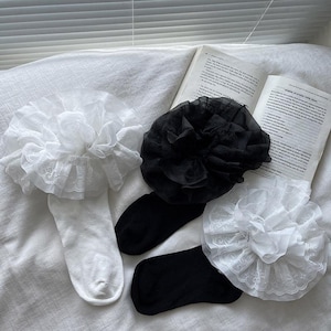 May include: Two pairs of socks with decorative floral ruffles. One pair is white with white ruffles and lace trim, the other is black with black ruffles. The socks are on a white surface with an open book in the background.
