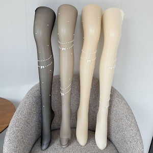 Könnte beinhalten: Vier Paar transparente Strümpfe in Schwarz, Beige und Creme, auf Schaufensterpuppen präsentiert. Jedes Strumpfpaar hat ein Perlen- und Schleifendesign. Die Strümpfe sind auf einem grau gepolsterten Stuhl angeordnet und zeigen die Details der Strümpfe.