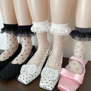May include: Five mannequin legs wearing sheer ankle socks with black or white polka dots and ruffled lace cuffs. The socks are paired with flat shoes, including black textured flats, white polka dot flats with a bow, and pink satin flats with a bow.