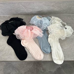 May include: Assortment of ruffled socks in various colors, including black, pink, blue, and white. Each sock features a delicate lace trim at the top. The socks are arranged on a light gray surface, showcasing their textures and designs.