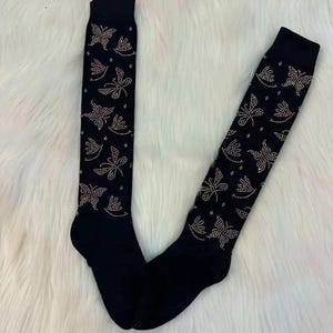 May include: A pair of black knee-high socks with a repeating pattern of butterflies and floral designs. The butterflies and flowers are embellished with small, sparkling rhinestones. The socks are displayed on a white, fluffy surface.