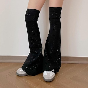 Puede incluir: Calentadores de piernas negros y brillantes con la parte inferior acampanada. Los calentadores de piernas se usan sobre zapatillas blancas con suelas grises. El fondo es una pared blanca lisa y un suelo de madera.