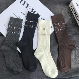 以下が含まれることがあります： チャコールグレー、ブラック、クリーム、ブラウンの4足のリブ編みソックス。各ソックスの足首上部には、フェイクパールと透明なラインストーンが2本の水平なライン状に埋め込まれており、直線的なデザインが施されています。