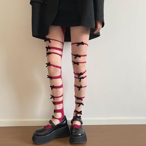 May include: Burgundy-colored thigh-high stockings with black bows. The stockings are paired with black platform Mary Jane shoes. The bows are evenly spaced along the red bands, creating a unique and stylish look. The outfit is completed with a black blazer and skirt.