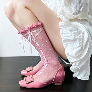 Peut inclure: Bottes roses montantes au genou avec un bout carré et un petit talon. Les bottes présentent une finition brillante, une boucle dorée et un motif en dentelle sur la partie supérieure. Des rubans blancs et des fausses perles ajoutent des détails décoratifs.