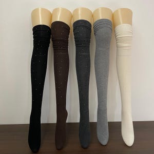 以下が含まれることがあります： 黒、茶色、濃いグレー、ライトグレー、オフホワイトのオーバーニーソックス5足。各靴下には、小さく散りばめられたキラキラ光る装飾が施されています。マネキンの脚に白い背景で靴下が展示されています。