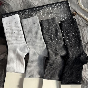May include: Four pairs of ribbed knit socks in white, light gray, dark gray, and black. Each sock is embellished with scattered, small, shiny rhinestones. The socks are displayed on white cardboard supports.