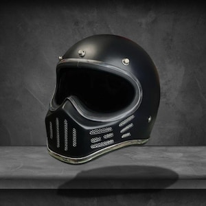 Może przedstawiać: Matowy czarny kask motocyklowy ze srebrnym wykończeniem wokół otworu na oczy i podbródka. Kask ma styl vintage z wentylowaną osłoną podbródka i gładką, zaokrągloną górą. Zaprojektowany z myślą o bezpieczeństwie i stylu.