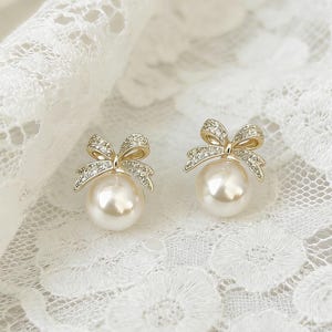 May include: A pair of pearl stud earrings with a bow design. The bows are encrusted with small, sparkling crystals. The earrings have a gold-colored metal setting and a large, round pearl. The text "Lizperry198929 Earrings" is at the bottom.