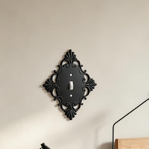 May include: A black diamond-shaped light switch cover plate featuring an ornate, scrollwork border with decorative fan-like accents at the top and bottom. The plate is mounted on a wall and includes a single white toggle switch in the center.
