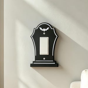 Könnte beinhalten: Eine schwarze, grabsteinförmige Lichtschalterabdeckung mit weißer Umrandung und einer kleinen weißen Fledermausgrafik an der Oberseite. Die Abdeckung ist an einer Wand montiert und umschließt einen rechteckigen, weißen Wippschalter mit zwei Schrauben.