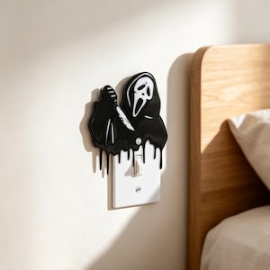 May include: A black and white light switch cover featuring a glossy, dripping design of the Ghostface mask character holding a knife. The cover is rectangular with a single toggle switch in the centre and two visible screws for mounting.