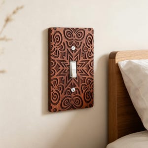 Puede incluir: Una placa de interruptor de luz rectangular de madera con un acabado marrón oscuro y un patrón geométrico simétrico profundamente grabado de líneas giratorias y formas de estrella. La placa está asegurada con dos tornillos plateados y tiene un interruptor blanco.
