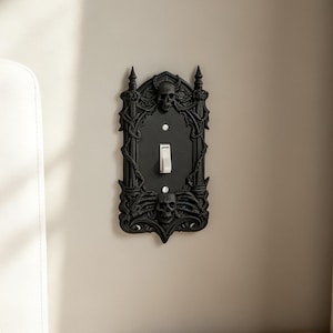 May include: A black rectangular light switch cover plate featuring an ornate gothic frame with two sculpted skulls at the top and bottom, intricate vine-like detailing, and two visible mounting screws surrounding a central white toggle switch.