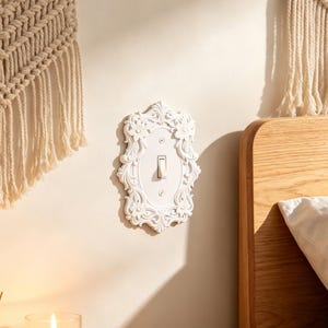 Può includere: Una placca per interruttore della luce bianca con un bordo ornamentale in rilievo raffigurante fiori e volute. La placca rettangolare presenta un interruttore a levetta centrale ed è fissata alla parete con due piccole viti bianche.