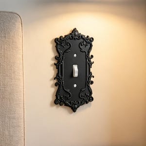 May include: A black light switch cover plate featuring an ornate, raised scrollwork border and a central white toggle switch. The rectangular plate is secured with two visible screws and displays intricate, symmetrical decorative detailing.