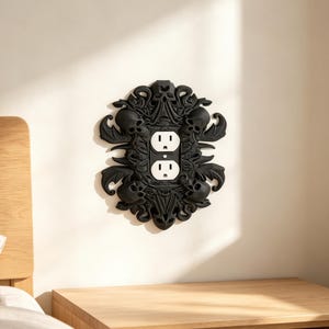 May include: A black decorative wall plate cover for a standard duplex electrical outlet. The cover features a detailed relief design of multiple skulls, swirling vines, and wing-like shapes surrounding the central white outlet receptacles.