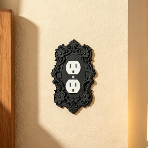 May include: A black electrical outlet wall plate featuring an ornate, raised relief design with floral motifs and scrolling patterns surrounding a standard white two-socket electrical receptacle with a central mounting screw.