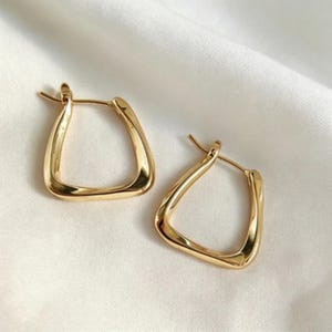 May include: A pair of gold-colored, square hoop earrings. The earrings have a smooth, polished finish and a unique, geometric shape. They are displayed on a white, textured surface, highlighting their reflective quality. The earrings are a stylish accessory.