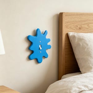 Op de afbeelding: Een felblauwe lichtschakelaar-afdekplaat met een onregelmatige, organische kloddervorm met afgeronde uitsteeksels. De afdekplaat is op een muur gemonteerd en omringt een standaard witte tuimelschakelaar die met twee kleine zilveren schroeven is bevestigd.
