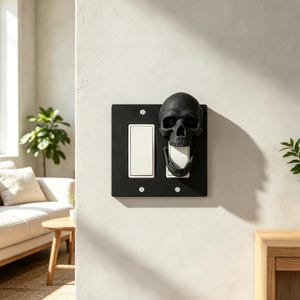 May include: A black double light switch wall plate featuring a three-dimensional skull sculpture attached to the right side. The skull has an open jaw that frames the right rocker switch. The plate is secured with four visible silver screws.