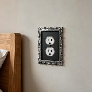 May include: A rectangular electrical wall socket cover featuring an ornate, silver-toned metallic frame with intricate scrollwork and decorative detailing. The centre plate is matte black and houses a standard white dual-socket electrical outlet.