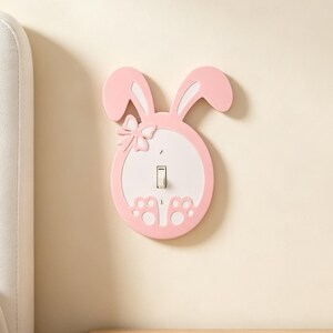 May include: A light pink and white light switch cover shaped like a rabbit. The cover features two long ears, a small bow detail on the left side, and two paw prints at the bottom. A single vertical toggle switch is centred in the white oval area.