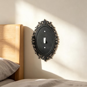 May include: An oval black light switch plate featuring an intricate, ornate filigree border. The central matte black panel contains a single white toggle switch and two small silver-toned mounting screws, creating a decorative wall fixture.