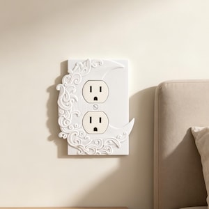 May include: A white rectangular electrical outlet wall plate featuring a raised, sculpted crescent moon design on the left side. The moon is decorated with intricate, swirling filigree patterns. The plate has two standard vertical outlet openings.