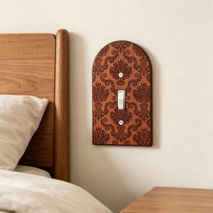 May include: An arched wooden light switch plate featuring a dark brown laser-engraved damask pattern. The plate is mounted on a wall and includes a central white toggle switch secured by two silver-colored screws at the top and bottom positions.