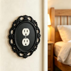 May include: A black oval wall plate cover featuring an ornate, decorative frame with scrollwork details. The plate is mounted over a standard white duplex electrical outlet, secured by a single central screw, and installed on a light-coloured wall.