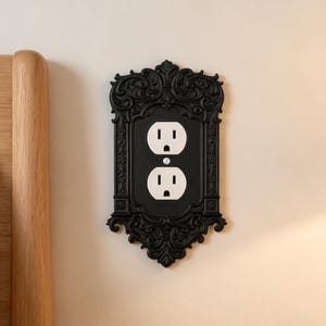 May include: A black electrical wall plate cover featuring an ornate, decorative frame with intricate scrollwork and floral detailing. The rectangular cover surrounds a standard white duplex electrical outlet with two vertical plug receptacles.