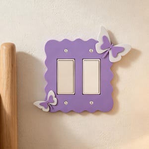 May include: A double light switch cover plate featuring a scalloped purple border and two white butterflies attached to the corners. The plate has two rectangular white rocker switches and is secured with four small silver-colored metal screws.