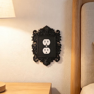May include: A black electrical wall plate cover featuring an ornate, scrollwork frame with intricate detailing surrounding a standard white duplex outlet. The rectangular cover is mounted flush against a plain, light-coloured wall surface.