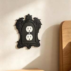 May include: A black wall plate cover for a standard duplex electrical outlet, featuring an ornate, arched frame with decorative wing-like motifs at the top and intricate scrollwork along the edges, surrounding a white two-socket power outlet.