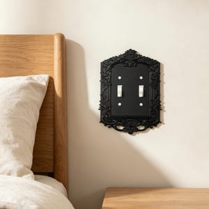 May include: A black double light switch cover plate featuring an ornate, raised relief frame with intricate floral and scrollwork detailing. The rectangular plate includes two vertical toggle switches positioned in the center with four screws.