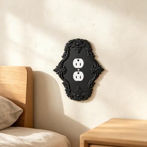 May include: A black wall plate cover featuring an ornate, decorative frame with intricate scrollwork and floral motifs surrounding a standard white dual electrical outlet. The rectangular plate has rounded, scalloped edges and a central screw.
