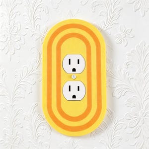 May include: An oval-shaped wall plate cover featuring a bright yellow and orange concentric ring pattern. The cover is designed for a standard duplex electrical outlet, with two white receptacles visible in the center and a single mounting screw.