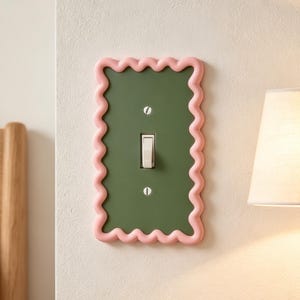 Può includere: Una placca rettangolare per interruttore della luce con un pannello centrale verde oliva e un bordo ondulato in rilievo di colore rosa chiaro. La placca è fissata con due viti argentate e presenta un interruttore a levetta bianco al centro.