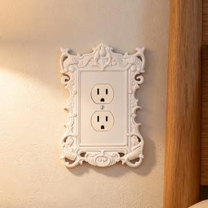 Może przedstawiać: Biała osłona gniazdka elektrycznego z ozdobną ramką, charakteryzującą się misternymi zawijasami i detalami w reliefie, otaczająca standardowe gniazdo z dwoma otworami. Prostokątna płytka maskująca jest zamontowana równo na teksturowanej ścianie.