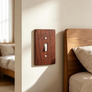 Puede incluir: Una placa rectangular de madera marrón oscuro para interruptor de luz con un diseño grabado a láser de dos manos que se extienden hacia un interruptor central blanco, rodeado de pequeñas estrellas y líneas punteadas radiantes.