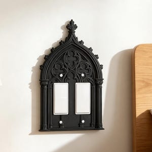 May include: A black double light switch cover plate featuring an intricate arched frame with pointed gothic-style detailing. The decorative plate surrounds two rectangular white rocker switches and is secured with four small white screws.