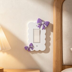 May include: A white rectangular light switch wall plate with a scalloped edge, featuring two purple bows attached to the top right and bottom left corners. The central rocker switch is beige and the plate is secured with two visible silver screws.