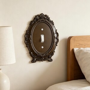 May include: An oval-shaped dark brown light switch cover plate featuring an ornate, raised relief border with scrollwork and floral details. The plate is mounted on a wall and includes a single white toggle switch secured by two silver screws.