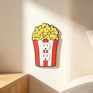 May include: A decorative wall plate cover shaped like a popcorn bucket with red and white vertical stripes and yellow popcorn at the top. The center features two white electrical outlet receptacles integrated into the popcorn bucket design.