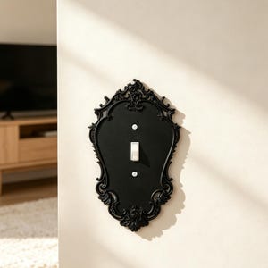 May include: A matte black light switch cover plate featuring an ornate, shield-shaped frame with intricate scrollwork detailing. The plate is mounted on a wall and includes a single white toggle switch secured by two visible silver-toned screws.