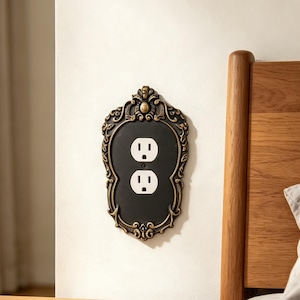 May include: A decorative wall plate cover for a standard duplex electrical outlet. The cover features a matte black centre panel surrounded by an ornate, raised bronze-toned frame with intricate scrollwork and leaf-like detailing at the top.