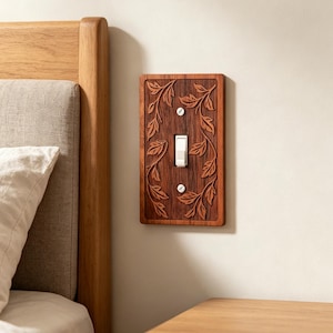 May include: A rectangular wooden light switch plate featuring a carved leaf pattern along the sides. The plate is dark brown with a visible wood grain texture and is secured to the wall with two white screws flanking a central white toggle switch.