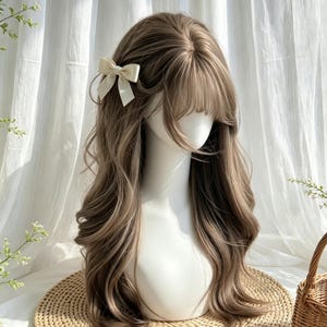 Puede incluir: Una peluca larga y ondulada de color marrón ceniza con capas suaves y flequillo desfilado, sobre una cabeza de maniquí blanca. El cabello tiene un flequillo lateral y está adornado con un pequeño lazo de tela color crema en el lado derecho.